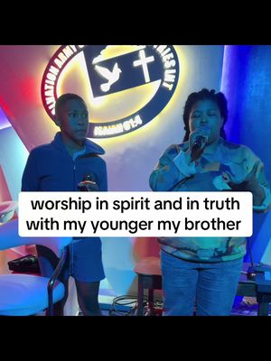 The cover image shows two individuals, presumably a woman and a man, on a stage setting. The woman is on the right, holding a microphone and singing; the man stands to her left, his gaze directed to the side. In the background, a circular logo with text and a cross is visible. The image is brightly lit, and a text overlay at the bottom reads 'worship in spirit and in truth with my younger my brother'.