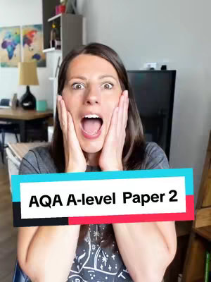 A woman with brown hair and a gray shirt is looking directly at the camera with a surprised expression, holding her hands up to her cheeks and mouth open. A text box at the bottom of the image reads, 'AQA A-level Paper 2'. The background includes a living room setting with a window, artwork, and furniture.