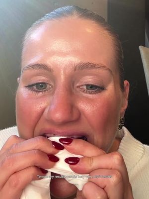 A close-up image of a woman with light skin, light-colored hair pulled back from her face, and dark red nail polish. She is holding a white paper in her mouth and appears to be biting down on it, with eyes wide and mouth ajar. Some areas of her skin are flushed red and in the background is a portion of a wall and a beige cushion.