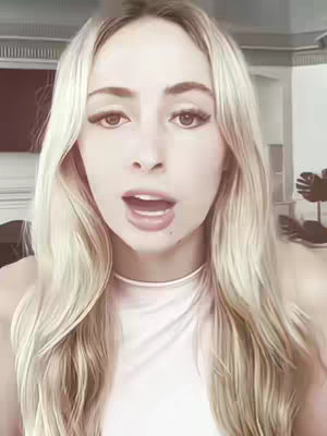 A head and shoulders shot of a woman with long blonde hair, looking directly at the camera with her mouth open as if speaking. She's wearing a white top and a thin necklace. The background is blurred.