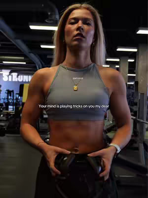 A woman with long blonde hair and a gray sports bra is standing in a gym, holding a black object. The background includes gym equipment and a sign that says "STRONG". Text overlayed on the image reads "Your mind is playing tricks on you my dear".
