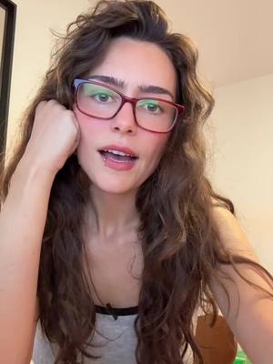 A young woman with long, wavy brown hair and glasses is shown in a close-up shot, expressing a thoughtful expression; she’s wearing a gray tank top and has a lip piercing. The background is a softly lit interior.