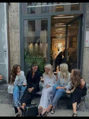 A group of five young women are seated casually on a ledge outside a building with glass doors. They appear to be waiting or taking a break, each holding a cup of something. The doorway behind them hints at an interior with tables and lighting.
