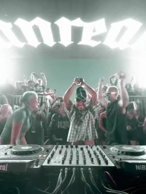 The cover image captures a lively music event, likely a concert or club night. Center stage, a DJ setup dominates the foreground with a mixer and two turntables, cables running below. Behind the DJ, a crowd of indistinct figures is packed tightly, with visible raised hands and enthusiastic reactions suggesting an energetic atmosphere. The background features a simple, flat wall, and the lighting appears to be from a combination of spotlights and the large illuminated text "umreo" hanging above, centered.