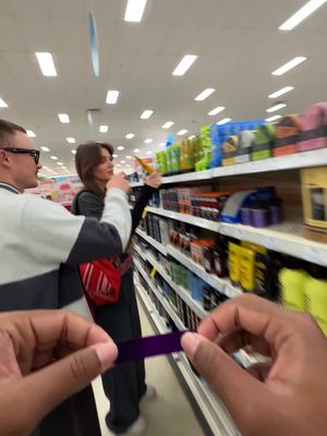 A selfie-style image taken from someone's perspective in a store. The foreground shows two hands holding a purple item, blurring the focus. In the mid-ground, two people are browsing shelves, one facing the camera and another looking at the shelves, lit by overhead lights and products. Shelves are stocked with colorful products.