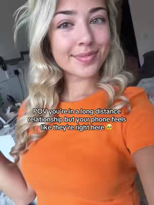 A selfie-style video cover image featuring a young woman with long, blonde hair and an orange top. She smiles and appears to be indoors. Text overlay reads: "POV you're in a long distance relationship but your phone feels like they're right here".