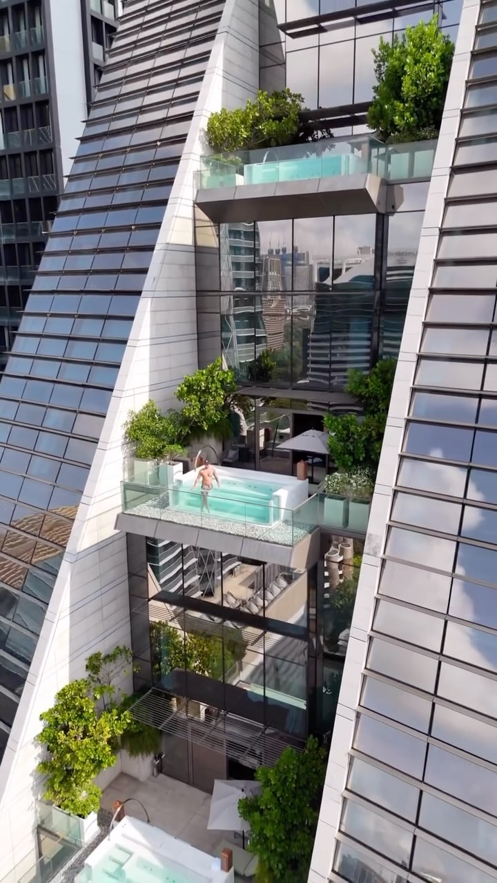 An elevated shot showcasing a modern skyscraper with a distinctive architectural design. The building features a unique, angular structure with floor-to-ceiling glass windows. The focus is on an open-air section of the building, where a man stands in a sleek, rectangular pool, surrounded by greenery. The composition is symmetrical, with the building's lines converging towards the top, creating a sense of height and grandeur.