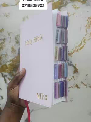 A person's hand holding a light pink Bible with a gold-colored "Holy Bible" text and NIV branding. Tabs are added to the side to separate the different books. In the background, a light colored, patterned design is visible. A price tag and phone number are in the top left.