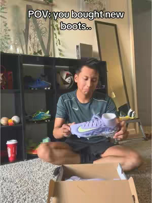 A man is sitting cross-legged on a carpet, holding a new pair of purple and yellow soccer cleats. The cleats are in an open shoe box. The background includes a shelf with soccer balls and other shoes, and a full-length mirror.