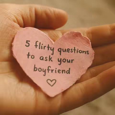 A hand holding a pink heart-shaped paper with handwritten text. The heart is slightly crumpled, suggesting it is handmade. The background is blurred, likely a close-up shot. The lighting is soft and warm.
