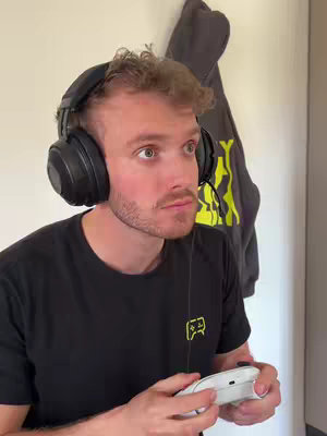 A young man wearing headphones and holding a white game controller looks upwards with a focused expression. The background includes a white wall, with a dark grey hoodie hanging on it.