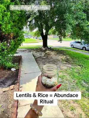 A person's hand holding a mason jar filled with lentils and rice in front of a house with a yard. The text overlay reads 'Lentils & Rice = Abundance Ritual'.