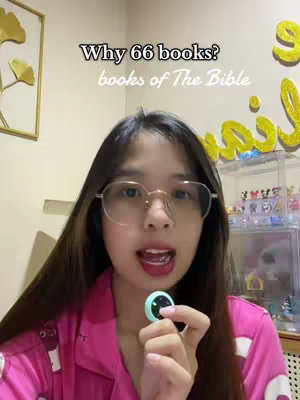 A woman with long dark hair, wearing glasses and a pink pajama top, is holding a microphone-shaped object. Text overlay reads 'Why 66 books? books of The Bible'. The background includes a wall and some decorations.