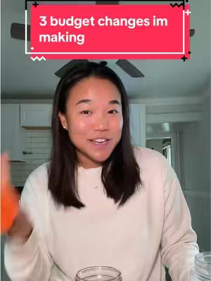 A woman is centered in the frame, smiling and speaking. Above her, a red banner reads '3 budget changes im making'. The background is a typical home interior, with a ceiling fan, lighting, and a glimpse of a kitchen.