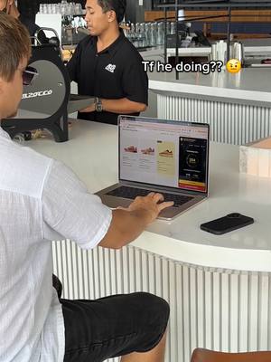 A person is sitting at a white counter with a laptop open in front of them, displaying a website. Another person is standing in the background, partially obscured, behind the counter. The laptop is the central focus, with a smartphone lying next to it. The scene is set indoors.