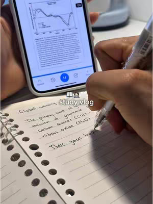 A hand writing notes on a spiral notebook, with a phone displaying text and graphics in the background. The layout comprises a close-up shot, emphasizing the writing process and the phone display.