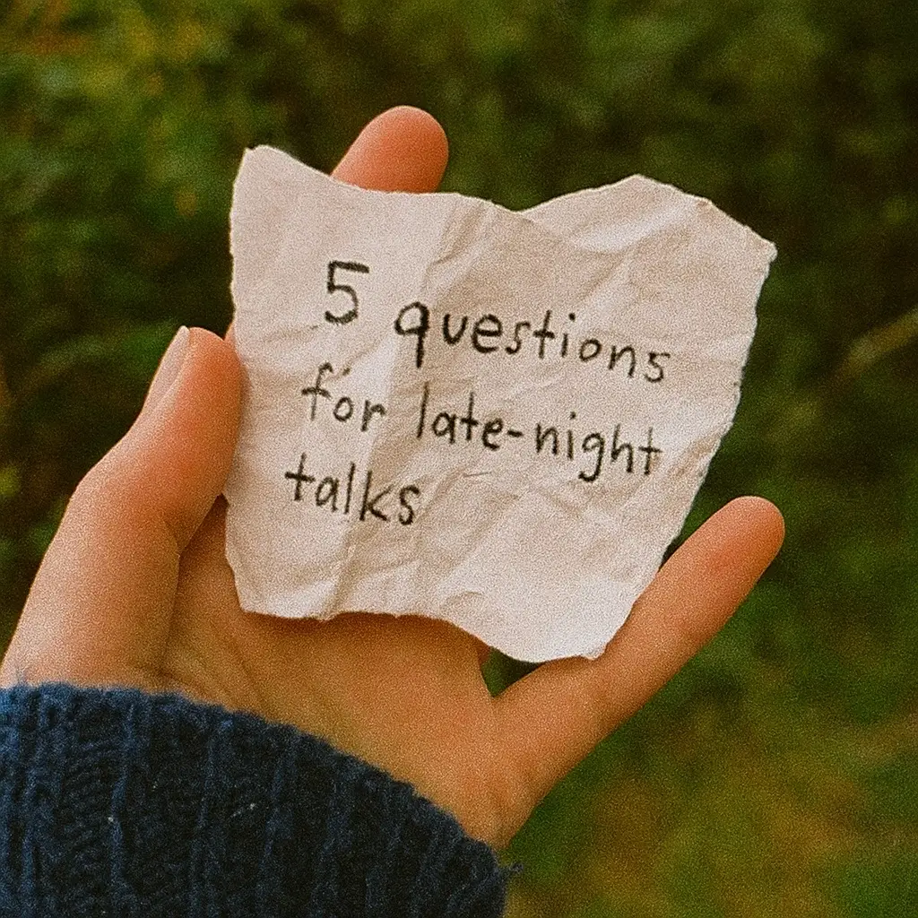 A hand holds a crumpled piece of paper with handwritten text against a blurry green background. The hand is fair-skinned and the sleeve of a dark blue sweater is visible. The background is out of focus greenery, suggesting a natural outdoor setting.