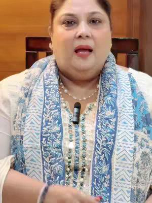 A medium shot features a woman with fair skin and short dark hair, positioned against a blurred background. She's wearing a white top and a patterned blue and white scarf that frames her face and shoulders. A pearl necklace with a small microphone is visible. She is looking upwards with her mouth open.