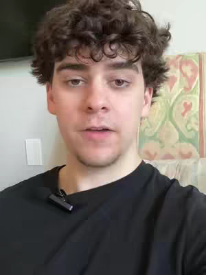 A young man with curly brown hair looks directly at the camera. He wears a black t-shirt, and a small microphone is visible clipped to his shirt. The background is blurred, but includes an off-white wall, a light switch, and a painting.