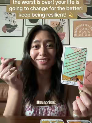 A woman with long dark hair smiles at the camera, holding a tarot card. Text overlays include inspirational messages. The background features framed artwork and a warm color palette.