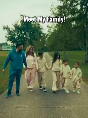 A family of seven walks towards the camera on a paved path. The father in a blue sweatsuit leads the way, followed by the mother and their children, all in matching outfits. Green trees and grass form the background.