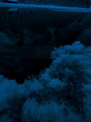 A dark, moody image dominated by shades of blue and black. The upper portion shows a dark, indistinct reflection of a road or structure. The lower half presents a turbulent, cloud-like form, resembling waves or billowing masses. The overall composition is abstract and atmospheric.