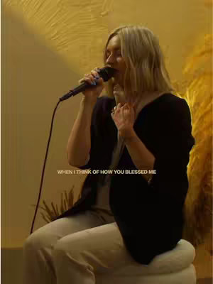 A woman with blonde hair is holding a microphone and singing. She wears a black top and cream-colored pants, sitting on a white stool. The background has a warm, golden tone.