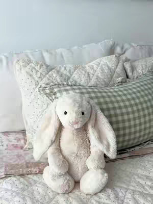 A plush white bunny sits centered on a bed. It has long, floppy ears and a pink nose. The bedding includes a floral quilt and patterned pillows in shades of white, light green, and pink, creating a soft and inviting atmosphere.
