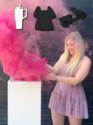 A woman stands with a pink smoke effect on a stand in front of a brick wall. There are objects above the woman's head: a white tumbler, a black dress, and a black scooter. The woman is wearing a pink dress and smiling.