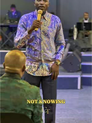 A man in a patterned shirt speaks into a microphone, set against a blurred background. A lower section of a person’s head and shoulders is visible in the foreground.