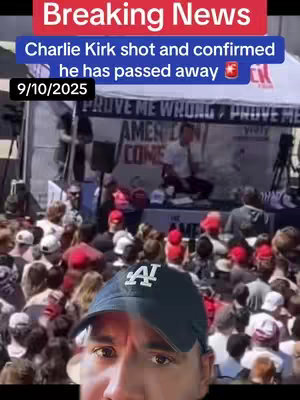 The cover image shows a man's face in the foreground, partially obscured by a baseball cap. Behind him, a large crowd of people are visible, gathered in what appears to be an outdoor event setting. The background includes tents, banners with text, and a stage area, suggesting a political or public speaking event. The top of the image features text overlays in red and blue boxes, providing a breaking news headline regarding Charlie Kirk. The overall composition is a mix of candid and staged elements, capturing a moment of public gathering while focusing on both the immediate foreground and a wider public scene.