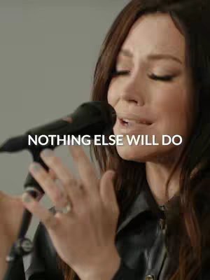 Close-up shot of a woman, likely a singer or worship leader, in a dimly lit setting. She is holding a microphone up to her mouth, eyes closed in a moment of emotional prayer or performance. The background is plain and out of focus, with text overlayed. The color scheme is muted, with warm skin tones in contrast with a black top and microphone.