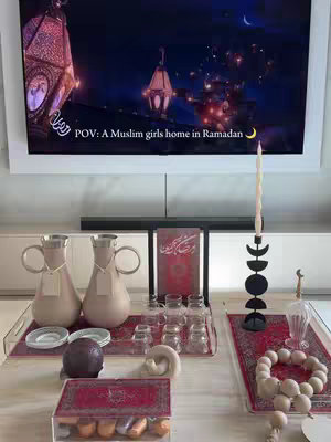 The image shows a well-lit living room setup, featuring a large TV screen displaying a digitally created scene of a Muslim home during Ramadan. The foreground showcases items arranged on a low table: decorative pitchers, glasses, a prayer book, candles, and decorative accents. The overall composition has a warm, inviting atmosphere, with a focus on traditional elements and soft lighting.