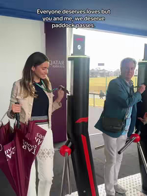 A woman and a man stand inside a building. The woman is on the left, holding two umbrellas and a lanyard around her neck. The man is to her right. A sign behind them says "Qatar Airways." Text above them reads "Everyone deserves loves but you and me, we deserve paddock passes."