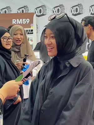 A woman with a black hijab and sunglasses perched on it smiles while speaking, holding a microphone as people surround her in a brightly lit room with signs and additional people in the background. Another woman is also holding a mic with her mouth open.