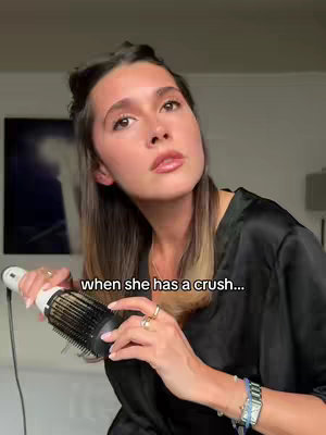 A woman with long hair angled to the right is holding a hair styling brush to style her hair. She is wearing a black robe. The hook that has been added to the cover image says 'when she has a crush...'.