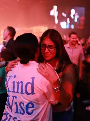 The cover image shows two women embracing with the one in the foreground wearing a white t-shirt with the words 'Kind Wise' printed on it. The woman in the foreground has dark hair and is hugging the woman behind her who has long dark hair and is wearing glasses. They both have tears in their eyes. The background is blurred, showing other people and a red-toned lighting.