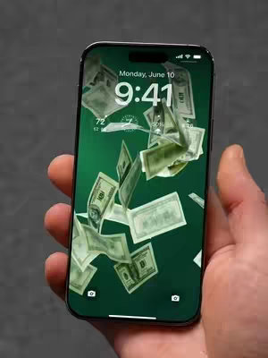 A hand holding a black iPhone displays a custom wallpaper of green background and American dollar bills floating in the air. The phone's screen displays the time at 9:41, and the date as Monday, June 10th. The device is held against a neutral gray background, and a light skin tone hand