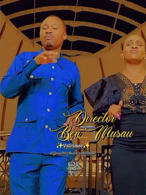 The cover image showcases two individuals, a man on the left and a woman partially visible on the right, against a beige background. The man, dressed in a blue suit, is the primary visual element, with his hands gesturing upwards. The woman wears a dark outfit. The text overlay, "Director Ben Musau" in a stylized script, is placed centrally, along with additional text elements. The overall composition features warm tones and a focus on the performers, suggesting a music video or performance-related context.