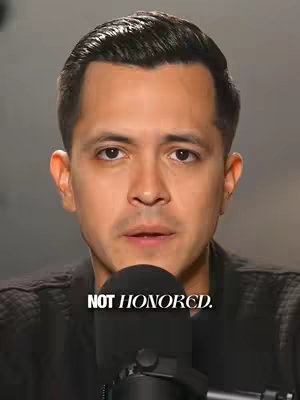 A medium shot of a man with short, dark hair facing the camera. He is wearing a black jacket and speaks into a black microphone. Text overlay 'NOT HONORED' is placed below the man.