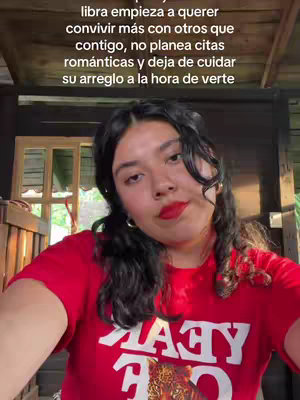A selfie-style shot featuring a young woman indoors. She wears a red t-shirt with white text, has dark curly hair, and is wearing red lipstick. The background shows a wooden structure and some greenery, adding to a casual setting. The text overlay adds context regarding astrological signs.