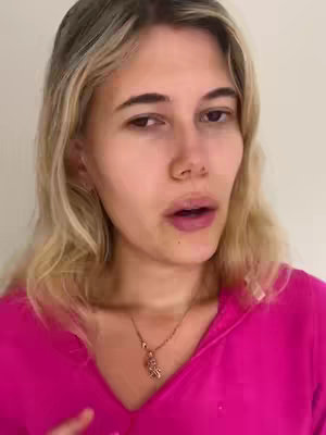 A woman with blonde hair and a hot pink top is centered in a close-up shot. She's looking slightly off-camera with a neutral expression. A gold necklace with a pendant hangs around her neck.