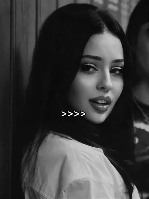 A grayscale photo features a woman with long, dark hair and fair skin. She's looking over her shoulder with a slight smile and red lipstick. Text overlayed on the image says "">>>>"