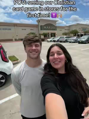 A selfie-style shot featuring a smiling couple standing in front of a retail store. The background includes the store's facade, other businesses, and a parking lot with cars. The sky is bright and clear, suggesting a sunny day. A text overlay at the top indicates the context of the scene.