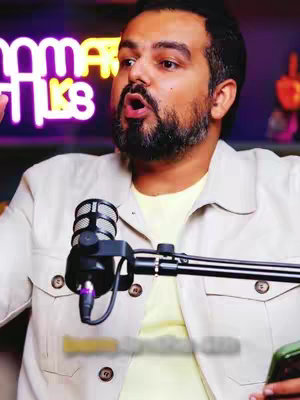A man is speaking into a microphone, with the name 'OMAR TALKS' in bright neon letters over his shoulder. He's wearing a beige button-up shirt over a light yellow undershirt. The background is a blurred gradient. There's a microphone in front of him.