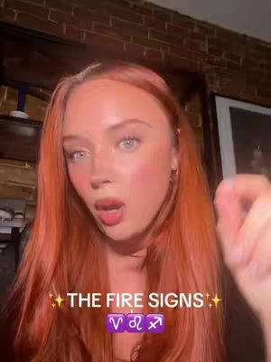 A young woman with long red hair and pink blush is looking directly at the camera with a surprised facial expression. The background includes exposed brick and wooden beams, suggesting an indoor setting. Text overlayed on the image reads 'THE FIRE SIGNS' along with the zodiac symbols for Aries, Leo, and Sagittarius, with animated sparkle effects.