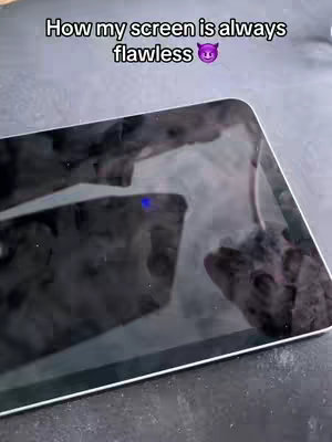 A close-up shot of a tablet screen on a dark surface. The screen reflects the surroundings, with text and a devil emoji overlaid. The screen itself appears to show surface blemishes. The background is dark, suggesting a low-key setting.
