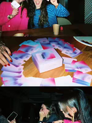 A multi-panel image showing scenes from a drinking game. The top panel features two young women smiling and holding cards. The middle panel focuses on the game cards spread out on a wooden table. The bottom panel shows two women in a dimly lit environment.