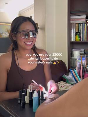 A woman wearing dental loupes is smiling and working on someone's nails. Nail polish bottles and a bottle of blue liquid are visible. The background includes a bookshelf and a painting.