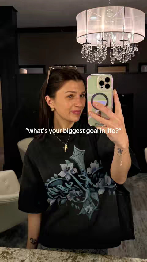 A woman takes a selfie in a room with a chandelier. She is wearing a black t-shirt with a floral design and a gold cross necklace. She is holding her phone up to take the picture. The background includes a dark wall and a glimpse of chairs.